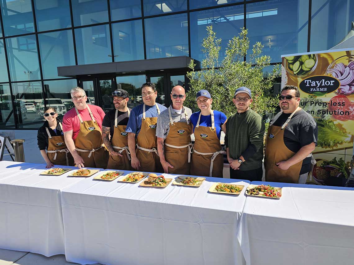 A group of eight people dressed in aprons and casual clothing stand behind a table displaying various plated dishes. They are posed outdoors in front of a modern glass building. A banner for Taylor Farms is visible in the background.